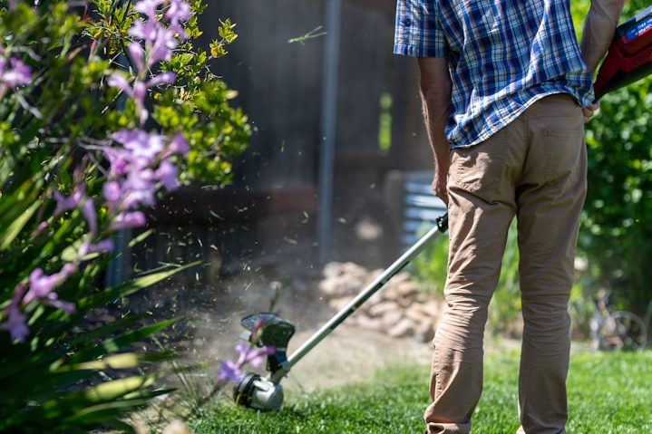 Mann mit Rasentrimmer schneidet Gras neben lila Blumen im Garten.