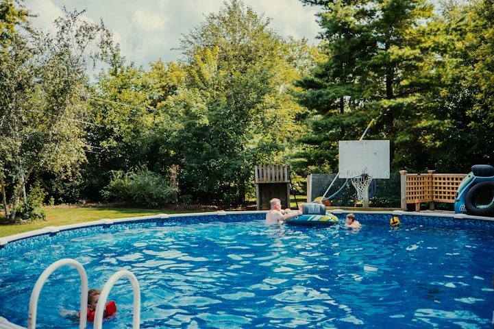 Kinder spielen in einem blauen Pool umgeben von Bäumen und grüner Landschaft.