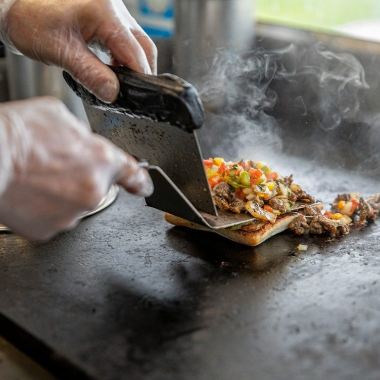 Plancha Eine Hand schneidet ein belegtes Sandwich mit Fleisch und Gemüse auf einer Grillplatte.