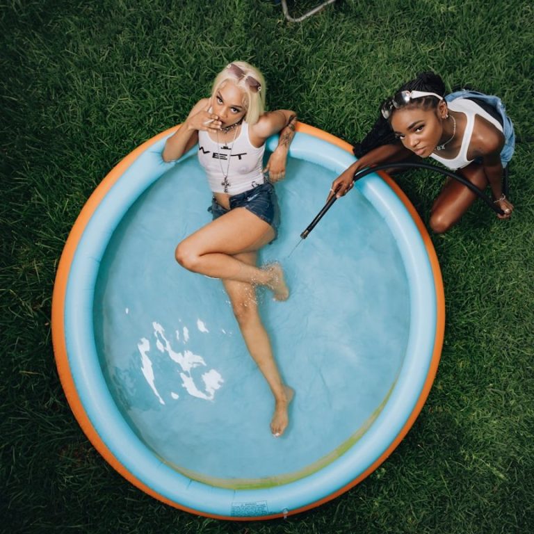 Prompt Set Pools Zwei Frauen, eine in einem Planschbecken, die andere mit einem Wasserspielzeug, auf grünem Gras.